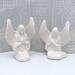 Set of 2 paintable ceramic angel statues sitting on a light blue table with an ecru lacy curtain behind them.